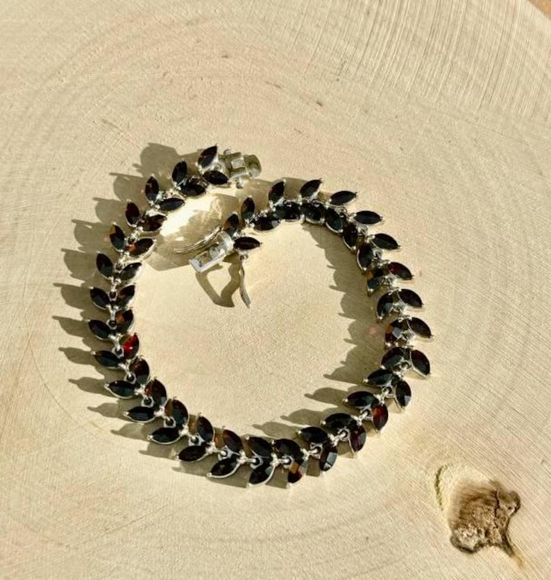May include: A silver bracelet featuring a row of dark red gemstone accents. The bracelet is made of small, connected links and has a clasp fastening.