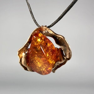 May include: A bronze pendant with a large amber stone set in a swirling, abstract design. The pendant is attached to a black cord.
