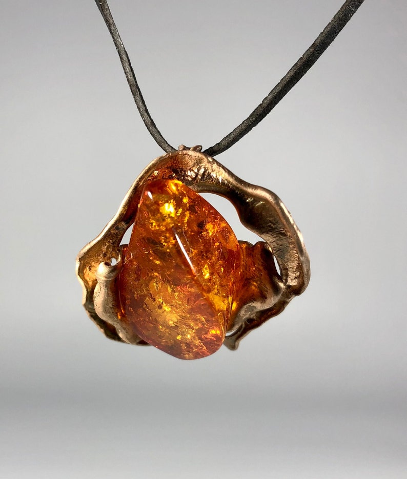May include: A bronze pendant with a large amber stone set in a swirling, abstract design. The pendant is attached to a black cord.