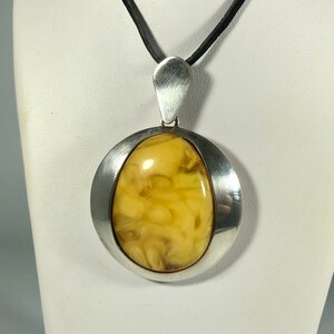 May include: A silver pendant with a large, oval-shaped amber stone. The amber is a light yellow color with brown swirls and inclusions. The pendant is on a black cord.
