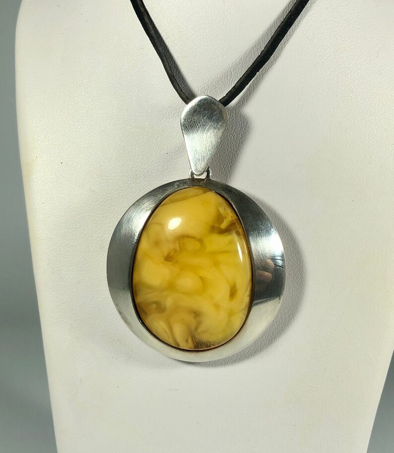 May include: A silver pendant with a large, oval-shaped amber stone. The amber is a light yellow color with brown swirls and inclusions. The pendant is on a black cord.