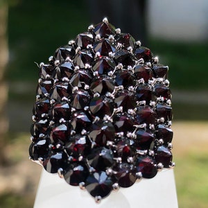 Pode incluir: Um anel grande de prata cravejado com um aglomerado de granadas vermelho-escuras. As granadas s&atilde;o facetadas e dispostas em um design pontiagudo em forma de cone.