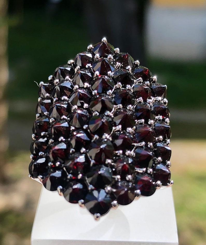 Pode incluir: Um anel grande de prata cravejado com um aglomerado de granadas vermelho-escuras. As granadas s&atilde;o facetadas e dispostas em um design pontiagudo em forma de cone.