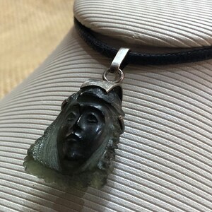 May include: A silver pendant necklace with a carved green stone featuring a face. The stone is a dark green color and the face is carved in detail. The pendant is on a black cord.