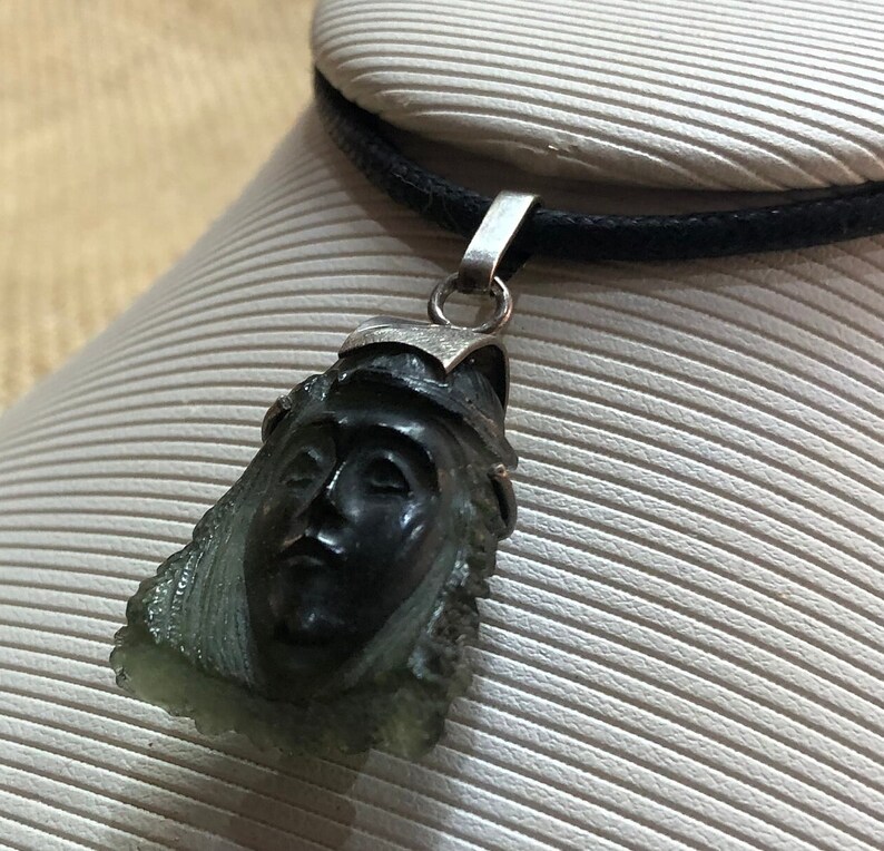 May include: A silver pendant necklace with a carved green stone featuring a face. The stone is a dark green color and the face is carved in detail. The pendant is on a black cord.