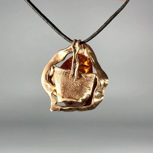 May include: A gold pendant necklace with a large amber stone set in a geometric frame. The necklace is on a black cord.
