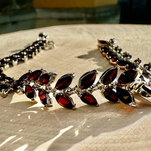 May include: A silver bracelet with a delicate design featuring a row of dark red gemstone leaves. The bracelet is made of metal and has a clasp closure.