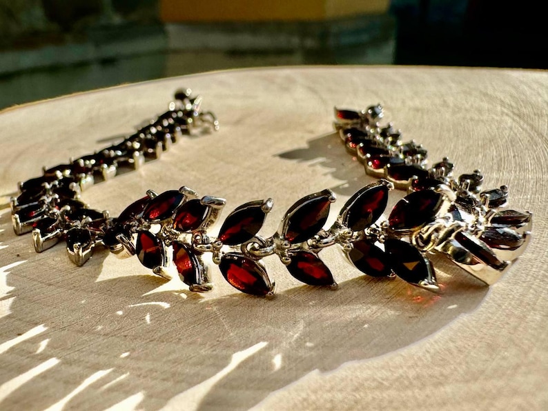 May include: A silver bracelet with a delicate design featuring a row of dark red gemstone leaves. The bracelet is made of metal and has a clasp closure.