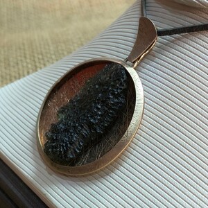 May include: A round silver pendant with a dark green and black stone inside. The pendant is on a black cord.