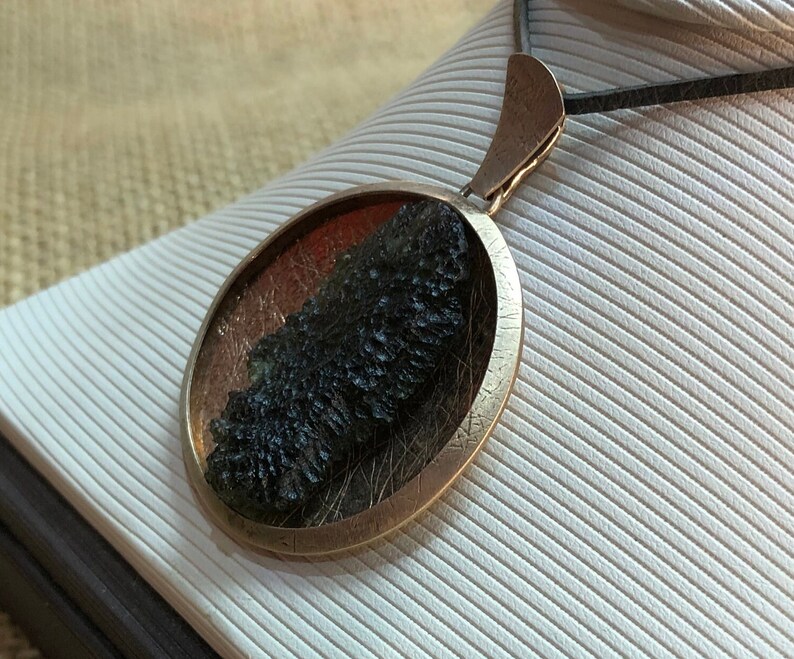 May include: A round silver pendant with a dark green and black stone inside. The pendant is on a black cord.