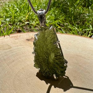 Puede incluir: Un colgante con una piedra de moldavita verde oliva rugosa. La piedra está sujeta por un engaste de metal plateado y cuelga de una cadena plateada. El colgante se exhibe sobre una superficie de madera con hierba verde en el fondo.