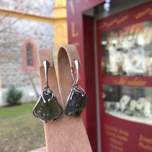 May include: A pair of silver earrings with green gemstone drops. The earrings are displayed on a brown velvet stand.