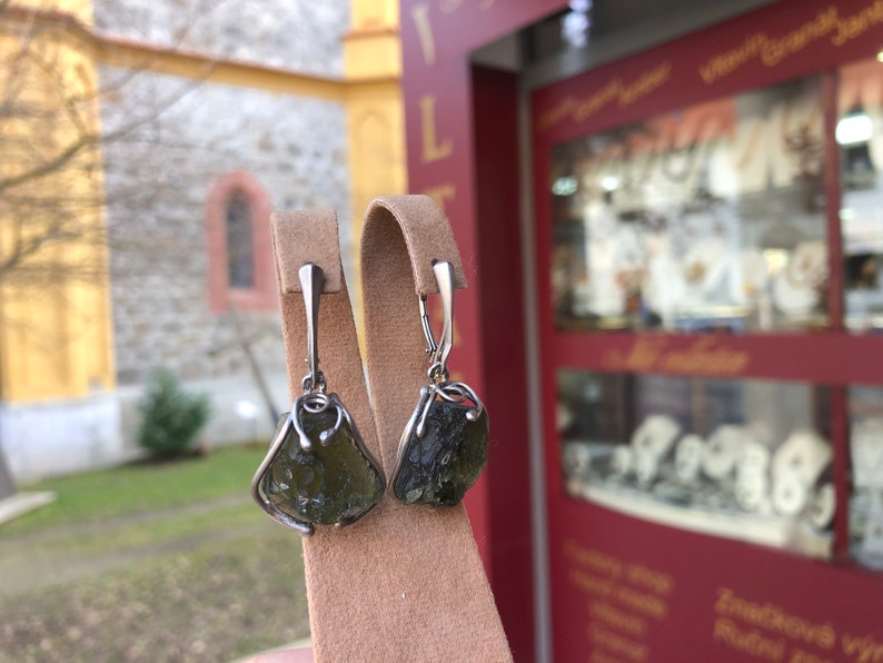 May include: A pair of silver earrings with green gemstone drops. The earrings are displayed on a brown velvet stand.