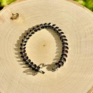 May include: A silver bracelet featuring a row of dark red marquise-shaped stones. The bracelet has a clasp fastening.