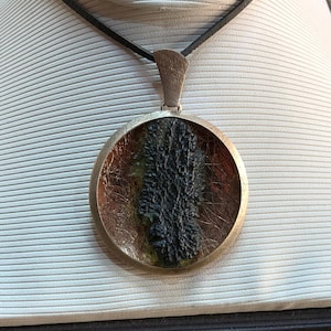 May include: A round silver pendant necklace with a black leather cord. The pendant features a black, green, and gold resin design.