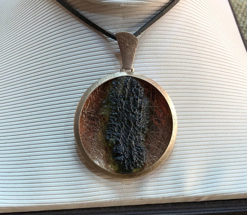 May include: A round silver pendant necklace with a black leather cord. The pendant features a black, green, and gold resin design.