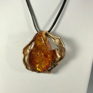 May include: A gold-toned pendant necklace with a large amber stone set in a textured, abstract metal frame. The necklace is on a black leather cord.