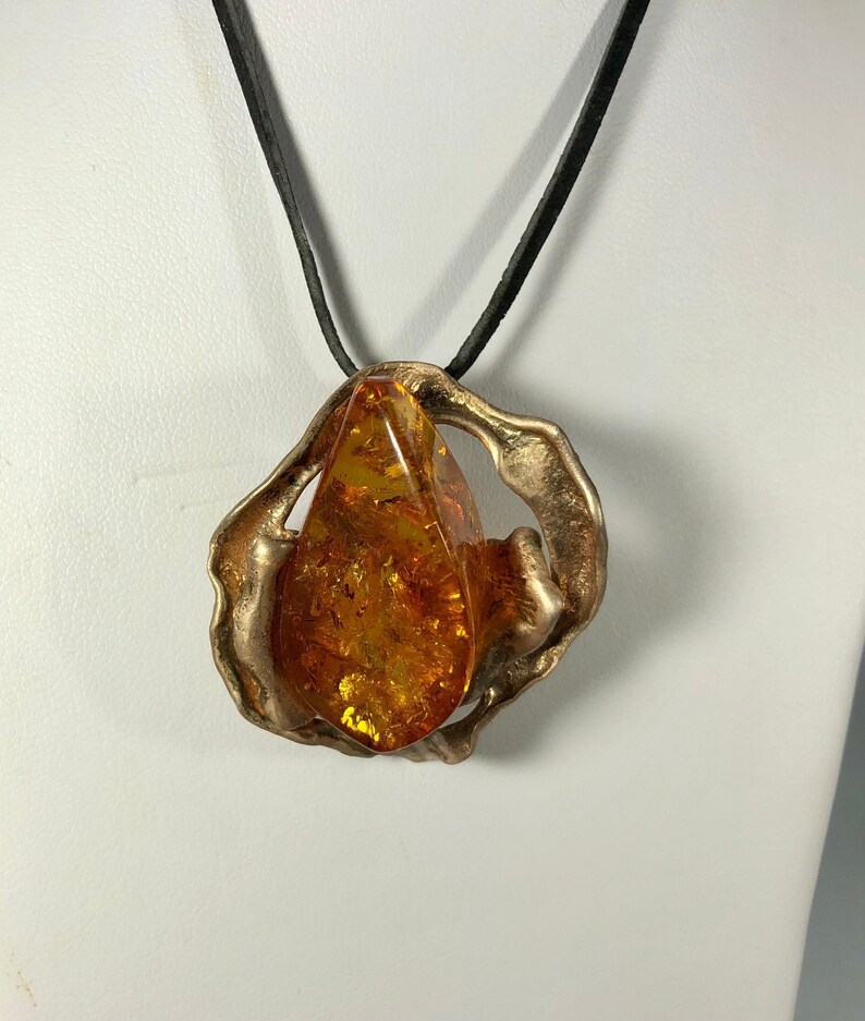 May include: A gold-toned pendant necklace with a large amber stone set in a textured, abstract metal frame. The necklace is on a black leather cord.