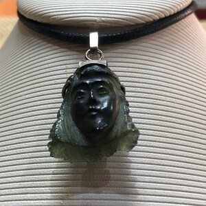May include: A silver pendant necklace with a carved green stone featuring a woman's face. The necklace is on a black cord.