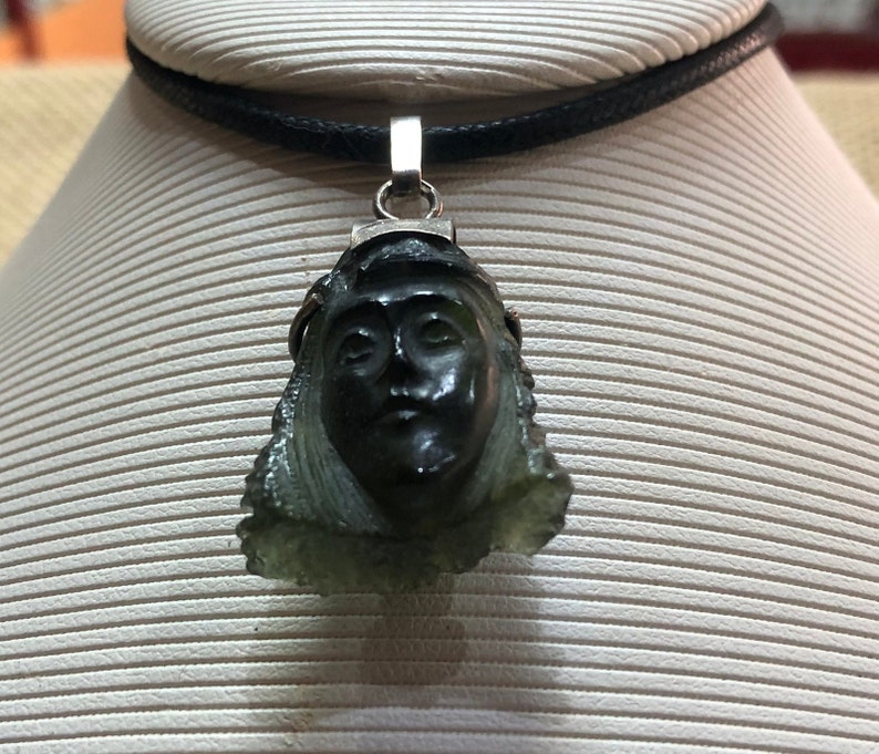 May include: A silver pendant necklace with a carved green stone featuring a woman's face. The necklace is on a black cord.