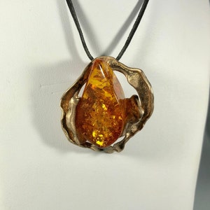 May include: A teardrop-shaped amber stone pendant set in a silver-toned, abstract, organic-shaped frame. The pendant is suspended from a black leather cord.