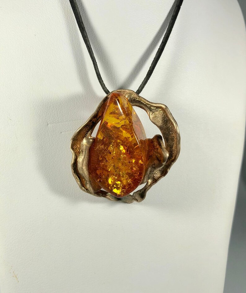 May include: A teardrop-shaped amber stone pendant set in a silver-toned, abstract, organic-shaped frame. The pendant is suspended from a black leather cord.
