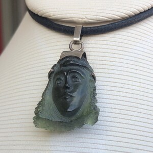May include: A green glass pendant necklace with a carved face, set in silver. The pendant is attached to a black leather cord.