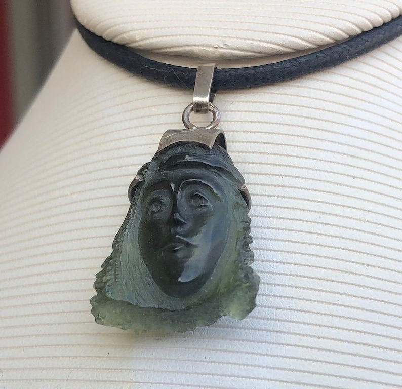 May include: A green glass pendant necklace with a carved face, set in silver. The pendant is attached to a black leather cord.