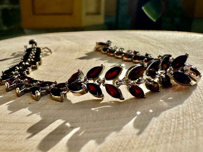 May include: A silver bracelet featuring a row of dark red marquise-shaped gemstones. The bracelet is crafted from a delicate chain and has a clasp fastening.