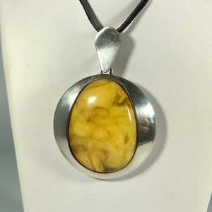 May include: A silver pendant with a large, oval-shaped amber stone. The amber is a light yellow color with brown swirls and inclusions. The pendant is suspended from a black cord.