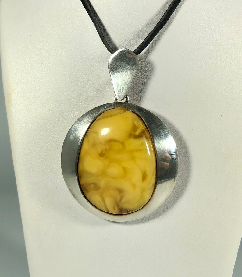 May include: A silver pendant with a large, oval-shaped amber stone. The amber is a light yellow color with brown swirls and inclusions. The pendant is suspended from a black cord.