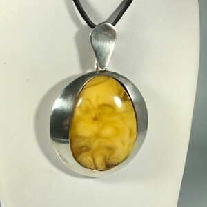 May include: A silver pendant with a large, oval-shaped amber stone. The amber is a light yellow color with brown streaks and swirls. The pendant is suspended from a black leather cord.