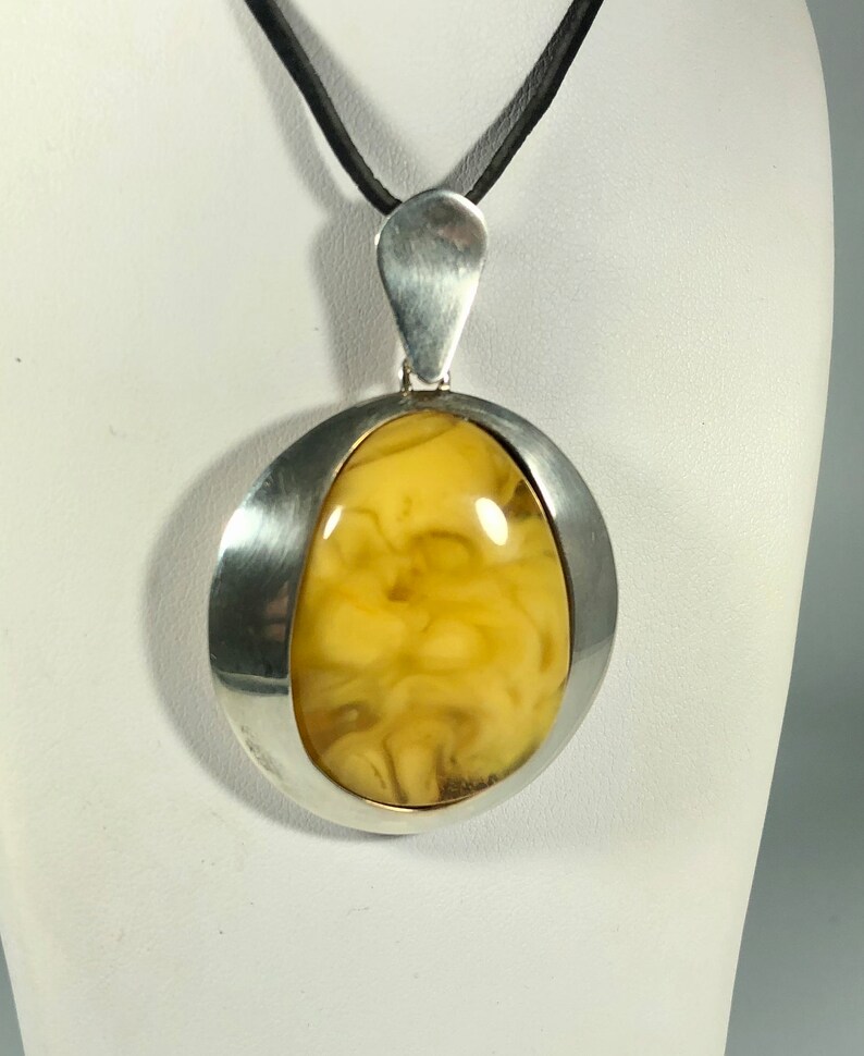 May include: A silver pendant with a large, oval-shaped amber stone. The amber is a light yellow color with brown streaks and swirls. The pendant is suspended from a black leather cord.