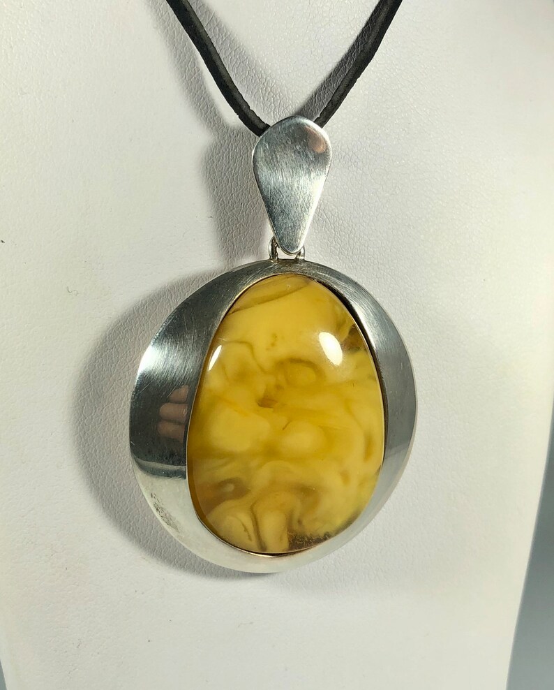 May include: A silver pendant with a large, oval-shaped amber stone. The pendant is suspended from a black leather cord.