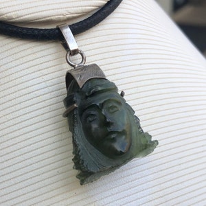 May include: A green jade pendant carved with a face, set on a black leather cord. The pendant is attached to a silver bail.