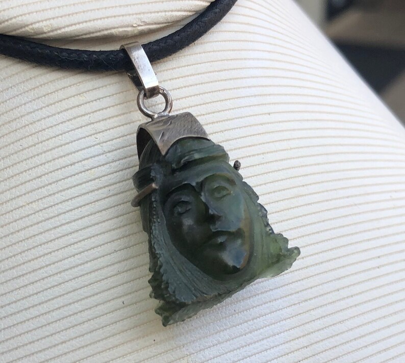 May include: A green jade pendant carved with a face, set on a black leather cord. The pendant is attached to a silver bail.