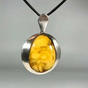 May include: A silver pendant with a yellow amber stone set in a smooth, oval-shaped frame. The pendant hangs from a black cord.