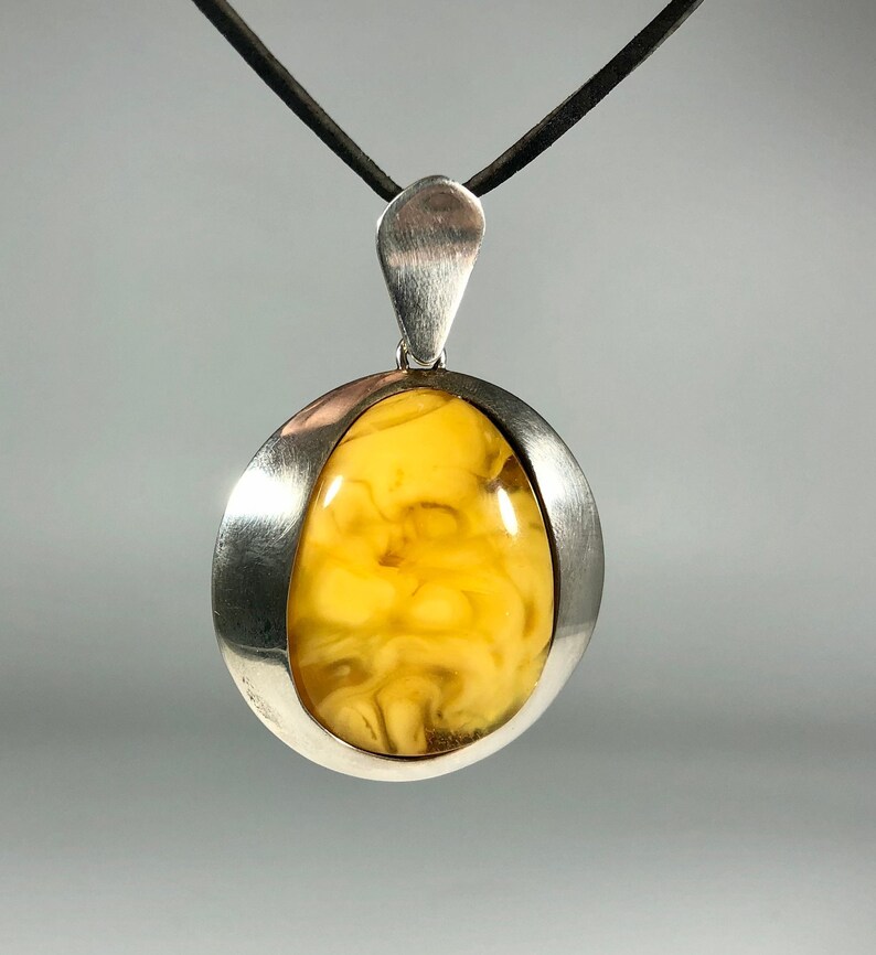 May include: A silver pendant with a yellow amber stone set in a smooth, oval-shaped frame. The pendant hangs from a black cord.