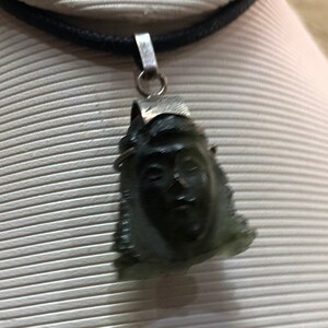 May include: A silver chain necklace with a dark green stone pendant carved with a face. The pendant is detailed with a silver crown.