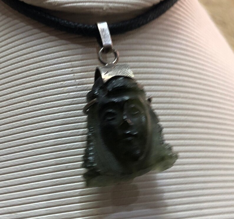May include: A silver chain necklace with a dark green stone pendant carved with a face. The pendant is detailed with a silver crown.