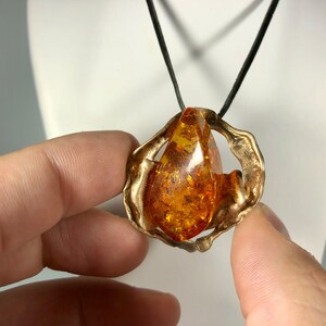 May include: A gold pendant necklace with a large amber teardrop stone set in a textured, abstract metal frame. The necklace is on a black cord.