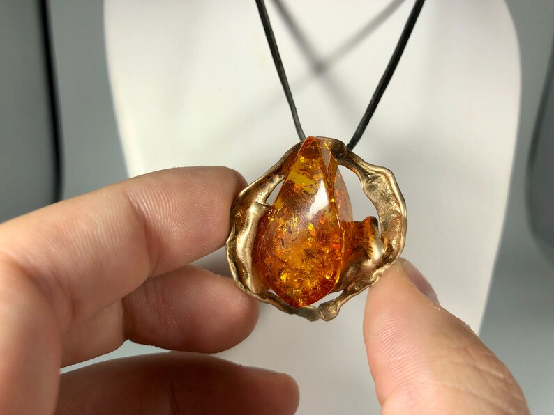 May include: A gold pendant necklace with a large amber teardrop stone set in a textured, abstract metal frame. The necklace is on a black cord.