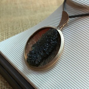 May include: A round, bronze pendant with a dark green, mossy stone inside. The pendant is on a black cord and is displayed on a white, textured surface.