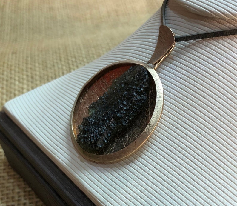 May include: A round, bronze pendant with a dark green, mossy stone inside. The pendant is on a black cord and is displayed on a white, textured surface.