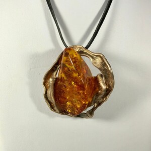 May include: A gold-toned pendant necklace with a large amber stone set in a unique, abstract design. The pendant is suspended from a black cord.