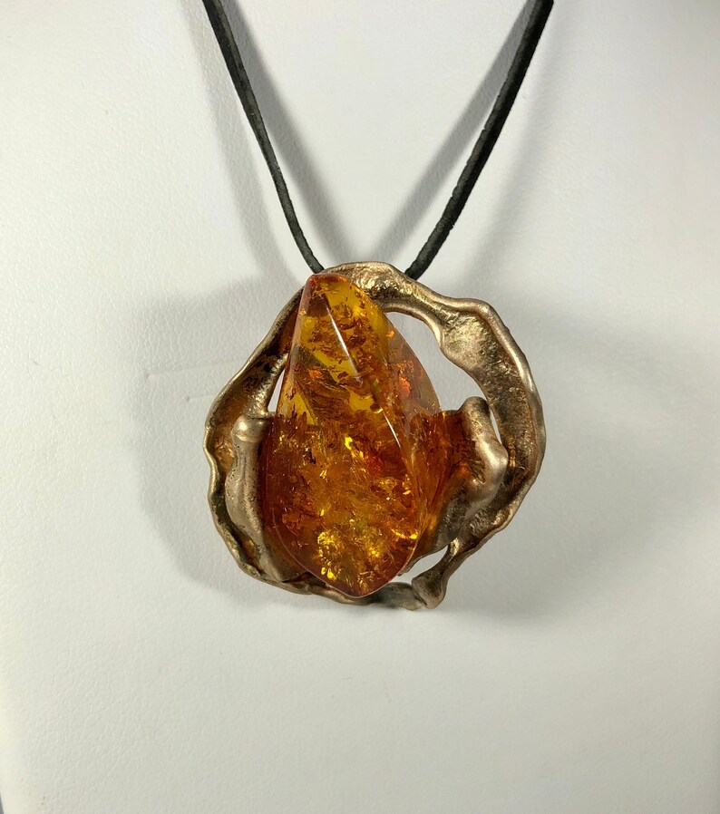 May include: A gold-toned pendant necklace with a large amber stone set in a unique, abstract design. The pendant is suspended from a black cord.
