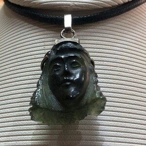 May include: A green stone pendant carved with a face, hanging from a black cord necklace. The pendant is attached to a silver bail.