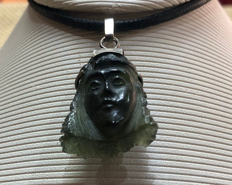 May include: A green stone pendant carved with a face, hanging from a black cord necklace. The pendant is attached to a silver bail.