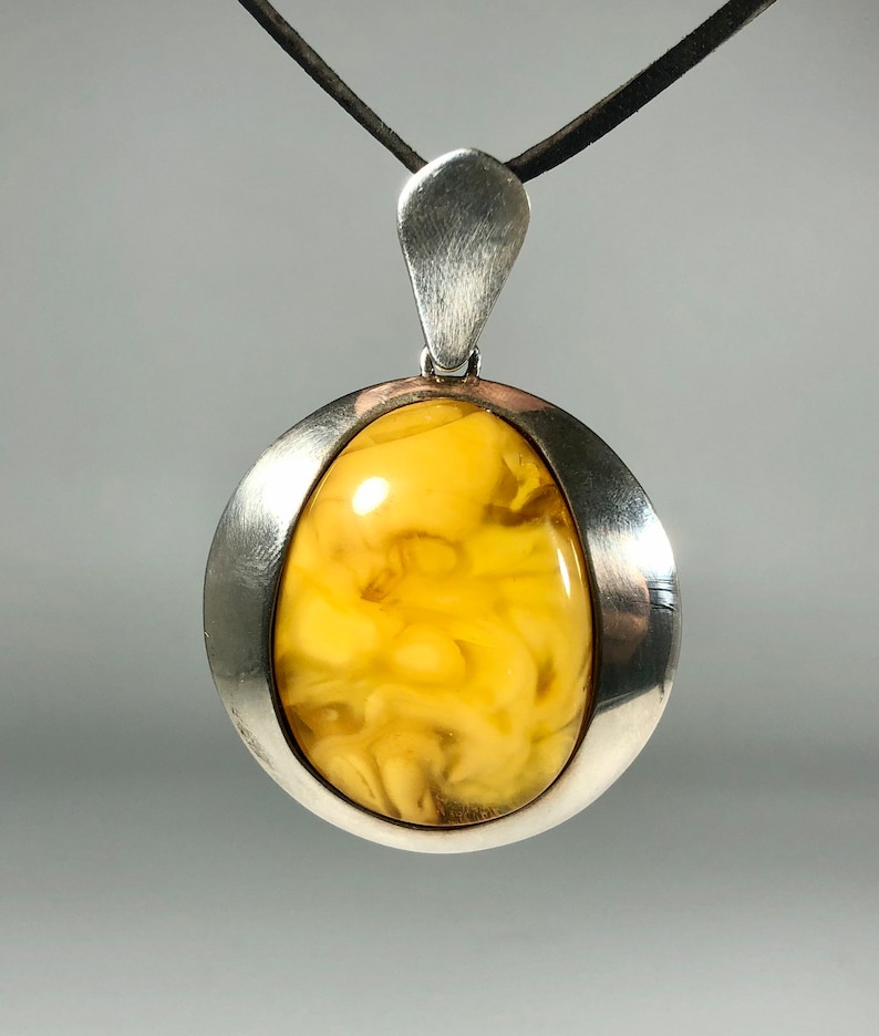 May include: A silver pendant with a large, oval-shaped amber stone. The amber is a light yellow color with swirls of darker yellow and brown. The pendant is on a black cord.