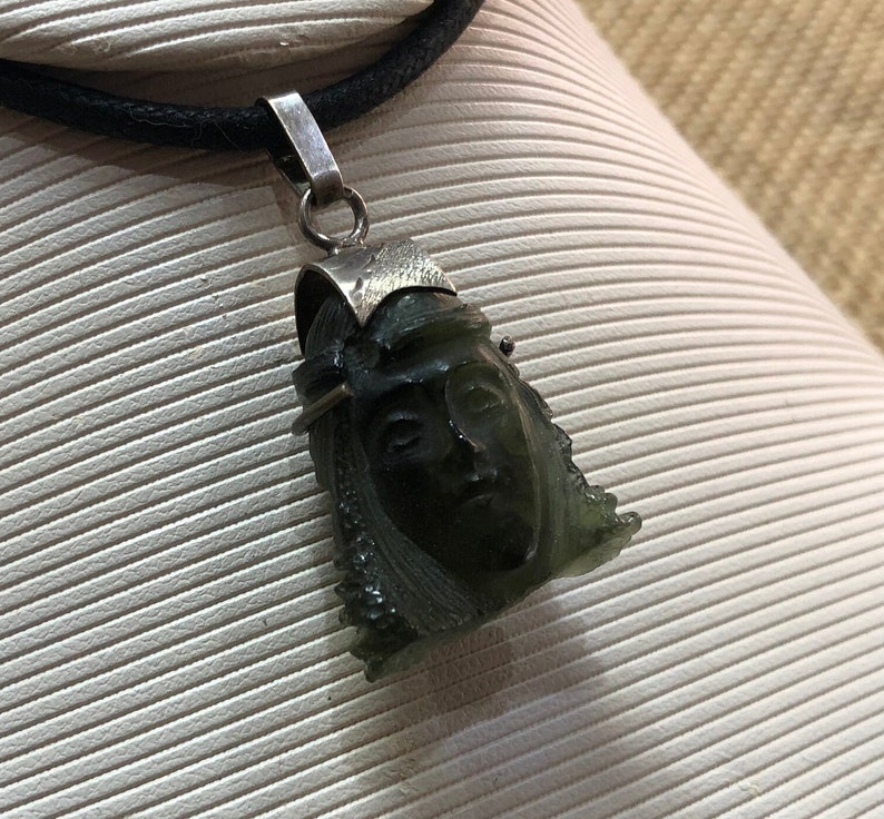 May include: A silver chain necklace with a green stone pendant carved with a face. The pendant is detailed with silver accents.